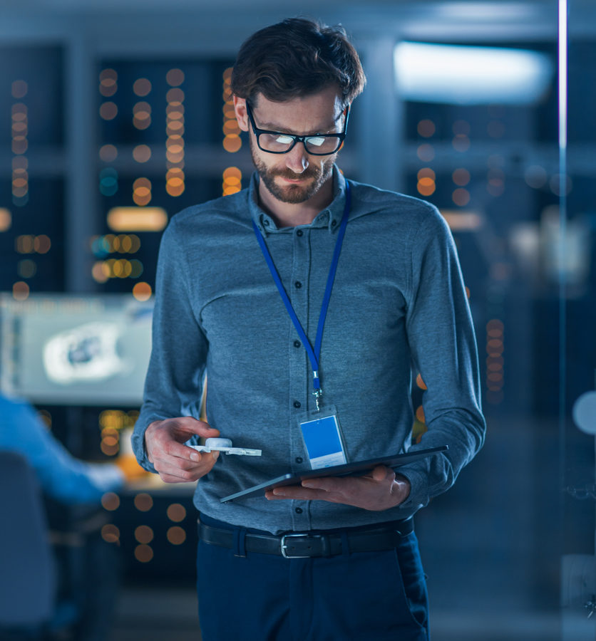 In Technology Research Facility: Chief Engineer Stands in the Middle of the Lab and Uses Tablet Computer. Team of Industrial Engineers, Developers Work on Engine Design Use Digital Whiteboard and Computers