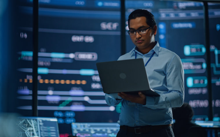 Young Multiethnic Male Government Employee Uses Laptop Computer in System Control Monitoring Center. In the Background His Coworkers at Their Workspaces with Many Displays Showing Technical Data.